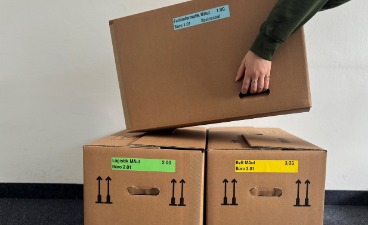 Zwei Umzugskartons stehen nebeneinander auf dem Boden und ein dritter wird auf diese abgestellt. Zu sehen sind nur die Arme un Hände.
